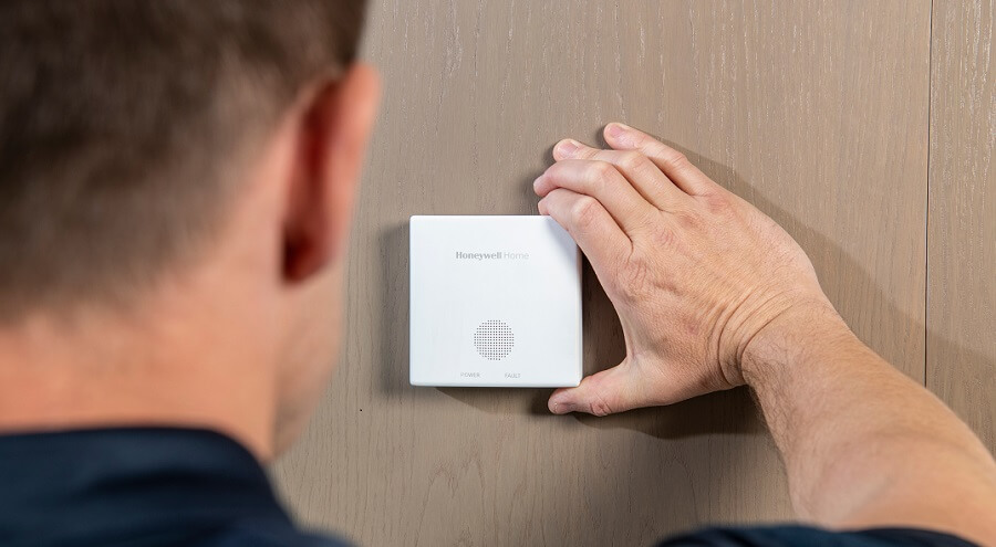 Muž přikládá detektor oxidu uhelnatého Honeywell Home ke stěně s dřevěným obkladem